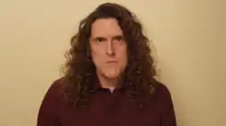 A man with long curly hair in a red dress shirt against a beige background.