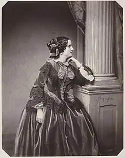 A black and white photograph of a woman wearing an elaborate dress. She is turned to the viewer's right, and is resting her chin on her left hand. Her left elbow is resting on the base of a column.
