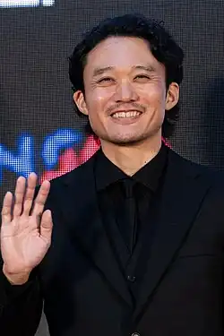 Fukunaga in three-quarter profile at press event wearing black suit and tie