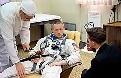 Armstrong, with short hair, partially reclining on a beige chair. He looks very serious. He is wearing a white space suit without a helmet or gloves. It has the U.S. flag on the left shoulder. Two hoses are attached. A technician dressed all in white is bending over him. A dark-haired, darkly dressed man has his back to us. He may be talking to Armstrong.