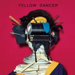 The album's cover art. Above a crimson red backdrop, everyday items like plates, cooking utensils, clothing, books, and a cat are arranged in a pile so that they resemble a human. Above the objects is the text "Yellow Dancer" in a standard font, all caps, and pure black.