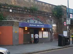 An entrance under a railway brick viaduct with a blue sign reading "GOLDHAWK ROAD STATION" in white letters and two women walking in front all under a grey sky