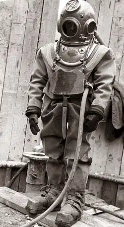 Standing figure of a diver clad in copper helmet, heavy canvas diving suit, with gloves, chest weight, and weighted boots