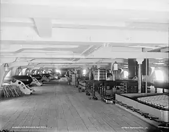 Black and white photograph of a wide wooden floor with low metal beams above and a row of cannons and gun ports on either side