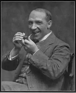 A black and white photo of a suited man, smiling whilst lighting a tobacco pipe.