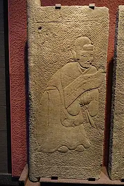 An Eastern Han carved stone tomb door showing a man carrying a shield, wearing a long robe with apron and a wuguan (武冠)/wubian (武弁) on jieze (介帻), stored in Sichuan Provincial Museum in Chengdu.
