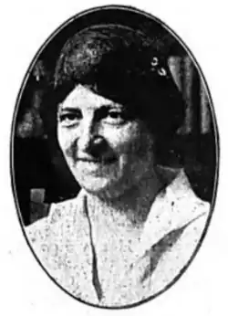 A smiling white woman with dar hair parted center, wearing a loose-fitting white blouse or dress with a wide collar, in an oval frame