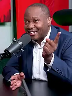 A man seated at a table and behind a microphone, wearing a navy blue suit and white collared shirt