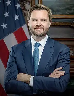 Official portrait of JD Vance, a middle-aged white man with dark hair and beard and light eyes, wearing a suit and tie, crossing his arms while standing in front of an American flag.