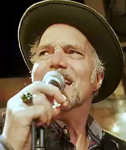 Caucasian man around 60, with light beard, singing and wearing a cowboy hat, with a ring on the hand holding the microphone.