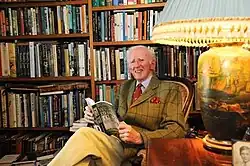 An older man in a book-lined study, wearing a tweed suit and reading a hardback book.