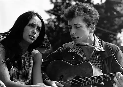 Dylan is seated, singing and playing guitar. Seated to his right is a woman gazing upward and singing with him.