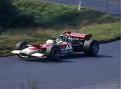 Side-view color photograph of Rindt racing a red and white Lotus Formula One car with a wing attached at the rear