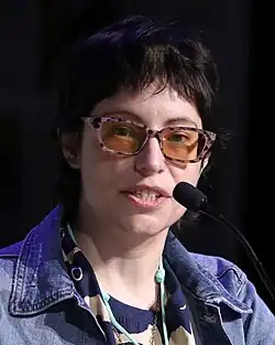 Photograph of Houser speaking into a microphone while looking towards the camera. She has short black hair and her glasses have orange tinted lenses.