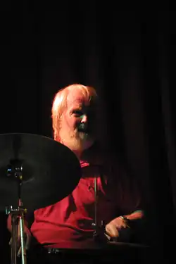 A bearded man in a red t-shirt behind a drum kit