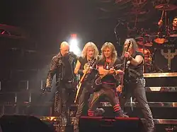 The band Judas Priest are onstage at a concert. From left to right are the singer, two electric guitarists, the bass player, and the drummer, who is seated behind a drumkit. The singer is wearing a black trenchcoat with metal studs.