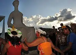 Indigenous people holding hands, two women in the foreground, surround a statue that represents Justice