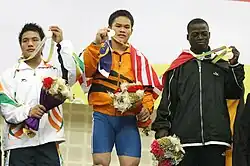 Aricco Jumitih lifting gold medal, flanked on left of image by Yukar Sibi (lifting silver medal) and on right by Charles Ssekyaaya (lifting bronze medal). All three medallists hold flower bouquets.