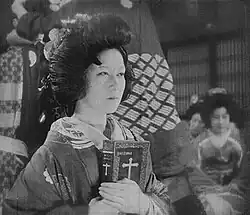 A film still in black and white, focused on a Japanese woman wearing a bouffant hair style, holding a large Bible in front of her chest