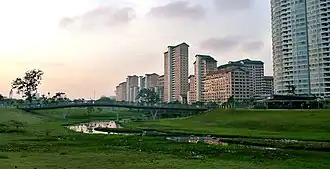 View of Bishan-Ang Mo Kio Park during sunset. The Kallang River is flowing around the grassy slopes dotted with a couple of trees, and there sits a bridge crossing above that. There are several housing buildings in the background.