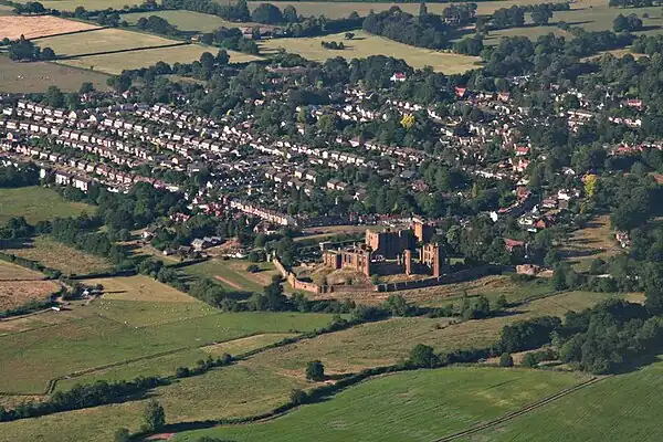 A red stone castle complex with a town on one side and open grassland on the other. While the castle is in ruins, from a distance large parts appear intact.
