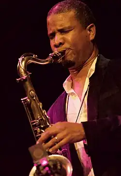 Close-up of a black man wearing a white shirt and a black vest playing the saxophone with his eyes closed.