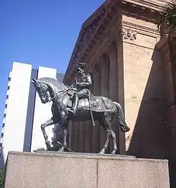 Equestrian statue in dark grey metal of George V in military dress uniform on a plinth of red granite outside a Classical building of red sandstone