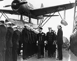 Midshot with a large single engined propeller warplane in background. A man with military ribbons covering his chest—the King (see caption)—accompanied by navy officers walks down a lines of sailors who stand rigidly at attention.