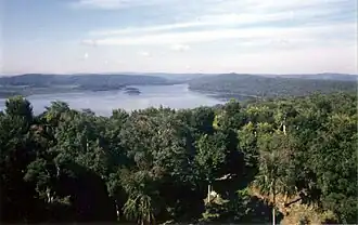 View across forest towards a lake surrounded by low, forested hills. Two small islands are set close to the far shore. There is a small clearing in the immediate foreground. On the left side of the far shore is a wide grassy region set against the forested hills.
