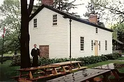 A white house in three-quarter perspective. In the foreground are several picnic benches. To the left is a tall tree with a man standing beside it. To the right, partly obsured by the house, is an old well.