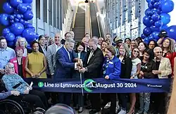 A group of people stand behind a wide ribbon reading "1 Line to Lynnwood [picture of train] August 30, 2024" with balloons in the background. Two people prepare to cut the ribbon with a pair of oversized scissors.
