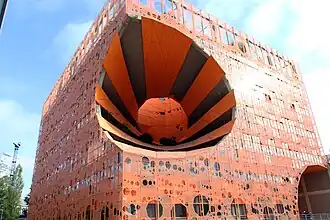 An orange multi-story building with a central open space and a perforated aluminum facade. It resembles a block of cheese with holes.