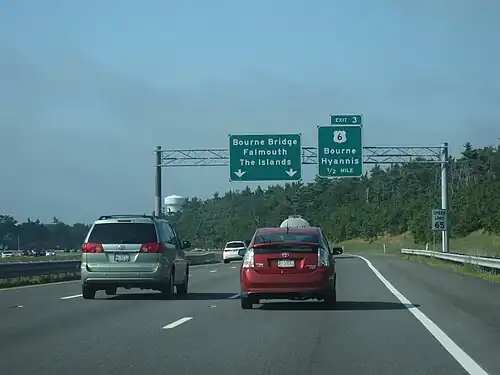 Ground-level view of three lanes of a divided expressway; two large green exit signs are visible in the distance, and the road is surrounded by dense forests.