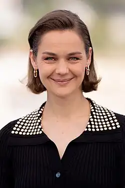 Luise Heyer wearing a dark top with white dots on turned-down collar, looking directly at camera with a broad smile