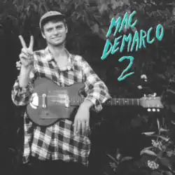 Grayscale picture of Mac DeMarco and his guitar. He is smiling and wearing a baseball cap.
