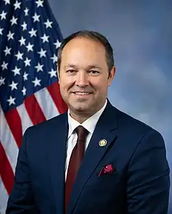 Stutzman smiling in front of the U.S. flag, wearing his House member pin, a black suit, white shirt, and red tie.