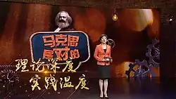 A woman stands on a television stage. Behind her, the image of Karl Marx is on the set's wall.