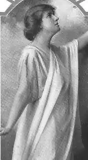A white woman wearing a white draped cloth, in a dance pose with both arms extending out of the frame of the image.