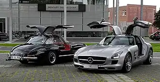 Two cars with open gullwing doors: an older black one and a gray one