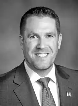 A monochrome portrait photo of a man with short hair, 5 o'clock shadow, wearing a suit coat and tie; he is looking and facing the center of the camera with a happy expression.