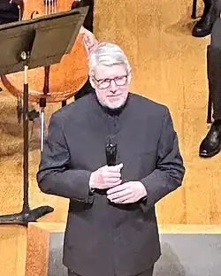 An older man in an all-black suit stands on a wooden stage with holding a wireless microphone in both hands.
