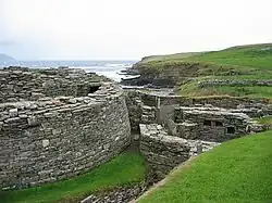 A semi-circular stone wall at left hints at the existence of a large and ancient building and to the right are the ruins of various other stone structures. In the background a low cliff divides a body of water from grassy fields.