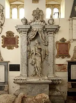 This image shows an elaborate baroque wall monument to the Hon. Margaret Watson (d.1713) by William Palmer at Rockingham, Northamptonshire. The monument features a dramatic architectural composition with a classical aedicule framed by fluted Corinthian columns supporting an entablature topped with heraldic achievements and decorative urns. The central figure of Margaret Watson stands in classical drapery within the columnar framework, whilst above her a large carved stone curtain appears to be drawn back, creating a theatrical canopy effect typical of late seventeenth and early eighteenth-century monument design. The curtain drapery demonstrates the period's fascination with trompe-l'oeil effects and references to contemporary funeral practices involving elaborate textile hangings. Below the figure is an inscribed memorial tablet, and the monument is surrounded by other wall memorials of various periods, including decorative cartouche-framed tablets. This monument exemplifies the grandeur and architectural sophistication of baroque commemorative sculpture, combining classical architectural elements with dynamic drapery to create a visually striking memorial composition.