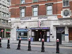 Across a street and behind black bollards is a brick building of at least two storeys. The ground floor is stone coloured and two people are standing in a dark entrance beneath a blue rectangular sign reading "MOORGATE STATION" in white. Above this, attached to be wall at 90 degrees is a white rectangular sign with the National Rail logo and London Underground roundel. Above this, below three windows, blue lettering reads "MOORGATE STATION"