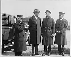Four men in naval jackets, three of them in caps and one in a top hat, standing to the right of a car, with two buildings in the background. Black and white photograph