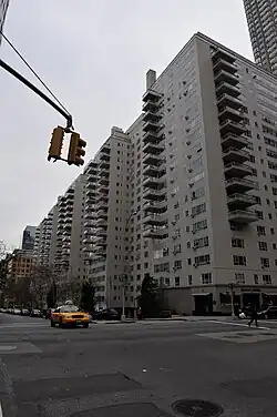 Manhattan House as seen from across the intersection of Third Avenue and 66th Street. The building contains a white brick facade with projecting glass balconies. There is a taxi on the road to the left.