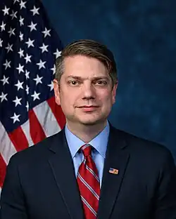 Official House portrait of Begich in front of the U.S. flag, wearing a black suit with American flag lapel pin, a blue shirt, and a checkered red and gray tie.