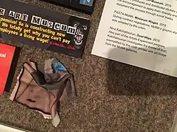 Crumpled microfiber dust wipe printed with an image of dust in a museum space and bearing the logo for the MoMA Artists Experiment program. An associated art label reads: "Also part of the Artists Experiment program, Katchadourian investigates the theme of dust, exemplified by this cloth printed with an image from the MoMA galleries."