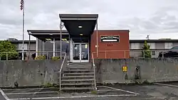 A covered walkway leads to glass double doors flanked on the left by floor-to-ceiling windows, and on the right by a red brick wall. On the brick wall is a rectangular sign, dark background, brass lettering/border reading "North Bend School District 13, We Are The Future, Administrative Offices".