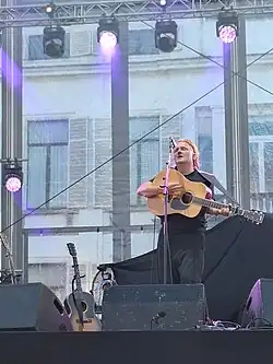 Joost Zweegers (Novastar) wearing a black t-shirt and dark pants, playing acoustic guitar and singing into a microphone onstage